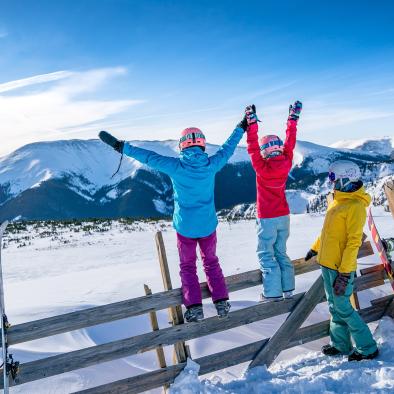 A couple young skiers raise their hands above their head as they look at an expansive view of snow-covered mountains